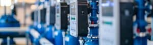 Close-up of several industrial control panels in a row, featuring digital displays with buttons. The panels are mounted on blue machinery, with a blurred background, suggesting a factory or mechanical setting.