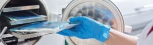 A person wearing blue gloves is placing a sealed blue packet into an open autoclave sterilizer. The metallic interior of the autoclave is visible, with racks for holding items. The setting appears to be a medical or laboratory environment.