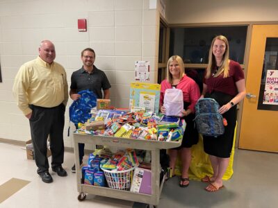 Four people stand by a cart filled with school supplies, including backpacks and notebooks. They are in a hallway, smiling at the camera.