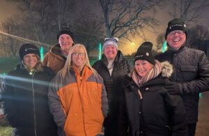 A group of six people standing outdoors at night, dressed warmly in coats and hats. There are trees and lights in the background, and the atmosphere appears festive and joyful. Everyone is smiling and huddled together.