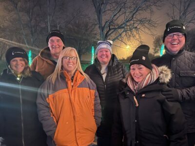 A group of six people standing outdoors at night, dressed warmly in coats and hats. There are trees and lights in the background, and the atmosphere appears festive and joyful. Everyone is smiling and huddled together.