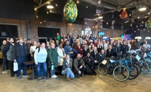 A large group of people is gathered in a spacious indoor area with exposed brick walls and bicycles lined up in the foreground. The ceiling has decorative, colorful hanging ornaments. Everyone is smiling and appears to be attending an event.