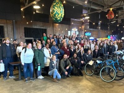 A large group of people is gathered in a spacious indoor area with exposed brick walls and bicycles lined up in the foreground. The ceiling has decorative, colorful hanging ornaments. Everyone is smiling and appears to be attending an event.