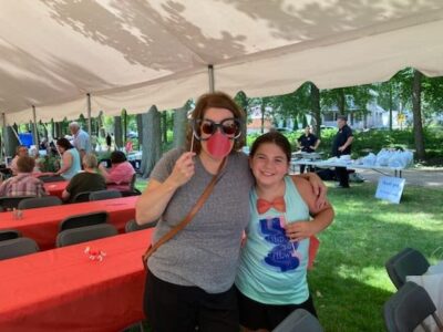 A woman and a girl smiling under a white tent at an outdoor gathering. The woman holds a paper apple prop. They stand near tables covered with red tablecloths, with more people and trees in the background.