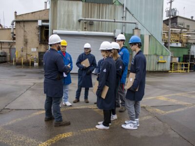 A group of people wearing hard hats and safety gear stand in a circle outside an industrial building. They are holding clipboards and appear to be in discussion, possibly during a safety briefing or site inspection. The area is paved and marked with yellow lines.