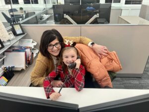 A woman and a young girl sit together at an office desk. The girl, wearing reindeer antlers, holds a phone receiver to her ear. The woman smiles warmly. The desk features office supplies and a computer monitor in the background.