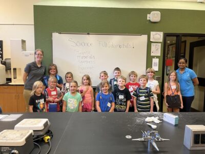 A group of young children and two adults pose in front of a whiteboard that reads "Science FUNdamentals!" The children are smiling and standing in a classroom setting with science equipment visible on the table.