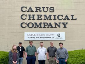 Six people standing in front of the Carus Chemical Company sign on a brick wall. They are all smiling and casually dressed. A smaller sign below mentions "Responsible Care" with an associated logo.