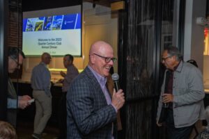 A man holding a microphone smiles while speaking at an event. People in the background are socializing near a screen displaying a presentation titled "Welcome to the 2023 Quarter Century Club Annual Dinner.