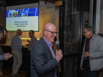 A man holding a microphone smiles while speaking at an event. People in the background are socializing near a screen displaying a presentation titled "Welcome to the 2023 Quarter Century Club Annual Dinner.