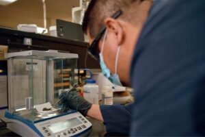 A person wearing safety glasses, a mask, and gloves operates a digital scale in a laboratory. They are carefully placing a small object on the scale inside a transparent enclosure. Various lab equipment is visible in the background.