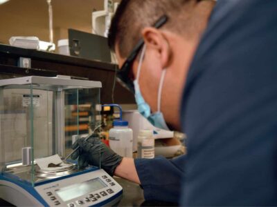 A person wearing safety glasses, a mask, and gloves operates a digital scale in a laboratory. They are carefully placing a small object on the scale inside a transparent enclosure. Various lab equipment is visible in the background.