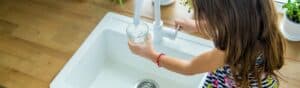 A child with long hair fills a glass with water from a white kitchen sink faucet. The countertop is wooden, and there are small potted plants nearby. The child is wearing a multicolored dress and a red bracelet.