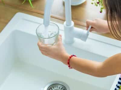 A young girl with long hair fills a glass with water from a kitchen faucet. She is wearing a colorful dress and a red bracelet. The countertop is wooden, and there are potted plants in the background.