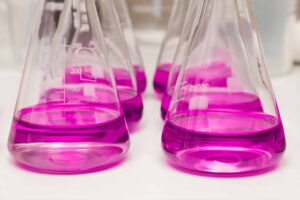 Several Erlenmeyer flasks filled with a bright pink liquid are arranged on a white surface. The flasks are transparent, allowing the liquid's hue to stand out vividly. Measurement markings are visible on the side of one of the flasks.