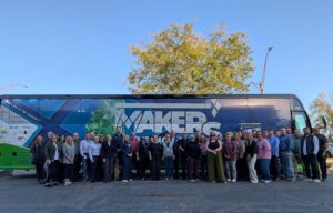 A large group of people stands in front of a bus with "MAKERS" on the side. The background shows a clear blue sky and a tree. Everyone is smiling and dressed in casual to business casual attire.