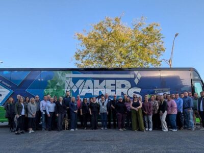 A large group of people stands in front of a bus with "MAKERS" on the side. The background shows a clear blue sky and a tree. Everyone is smiling and dressed in casual to business casual attire.