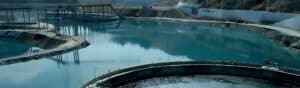 A large, circular water treatment facility with clear blue water is surrounded by a rocky landscape and other structural elements. The background shows a hilly area with some construction work visible.