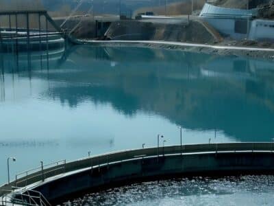 A panoramic view of a water treatment facility surrounded by hills. The facility features circular tanks and large pools reflecting the clear blue sky. Industrial structures and equipment are visible around the water bodies.