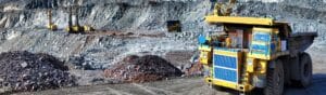 A large yellow mining truck is parked in an expansive open-pit mine. In the background, rock formations and smaller drilling machines are visible. Piles of extracted rock are scattered around the site under a clear sky.