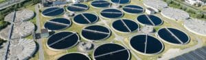 Aerial view of a wastewater treatment plant featuring numerous circular tanks arranged in a grid pattern. The facility is surrounded by greenery and a few buildings, with roads connecting the different sections.