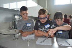 Three boys in a science lab, wearing safety goggles and name tags, engage in an experiment. They are focused on a task involving a beaker and tongs, with notebooks and lab equipment on the table.