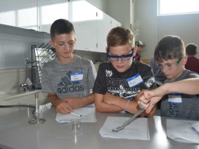 Three boys in a science lab, wearing safety goggles and name tags, engage in an experiment. They are focused on a task involving a beaker and tongs, with notebooks and lab equipment on the table.