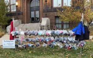 A community donation fence outside a brick building displays various winter clothing items such as coats, hats, and scarves in clear plastic bags. A sign reads, "The Giving Fence: Take what you need, leave the rest.