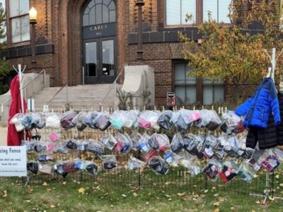 A community donation fence outside a brick building displays various winter clothing items such as coats, hats, and scarves in clear plastic bags. A sign reads, "The Giving Fence: Take what you need, leave the rest.