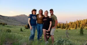 A group of four people stand smiling in a grassy field with rolling hills and pine trees in the background. One person wears a cowboy hat, and another has a knee brace. The sky is clear with a hint of sunset colors.