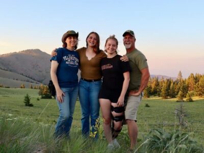 A group of four people stand smiling in a grassy field with rolling hills and pine trees in the background. One person wears a cowboy hat, and another has a knee brace. The sky is clear with a hint of sunset colors.