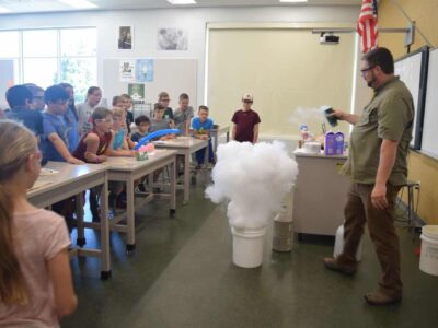 A teacher conducts a science experiment in a classroom, creating a large cloud of vapor from a bucket. Students watch enthusiastically from behind desks. The room has posters and a U.S. flag on the wall.