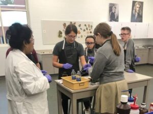 A group of students wearing gloves gather around a lab table with a teacher in a lab coat. They're examining items on the table. The room is equipped with lab materials, and botanical charts are visible on the wall behind them.