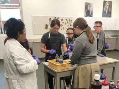 A group of students wearing gloves gather around a lab table with a teacher in a lab coat. They're examining items on the table. The room is equipped with lab materials, and botanical charts are visible on the wall behind them.