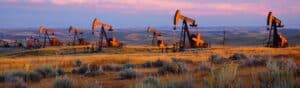 Oil pumps operating in a vast, rolling field during sunset. The landscape features dry grasses and sparse vegetation under a colorful sky with pink and purple hues. The machinery stands silhouetted against the setting sun.