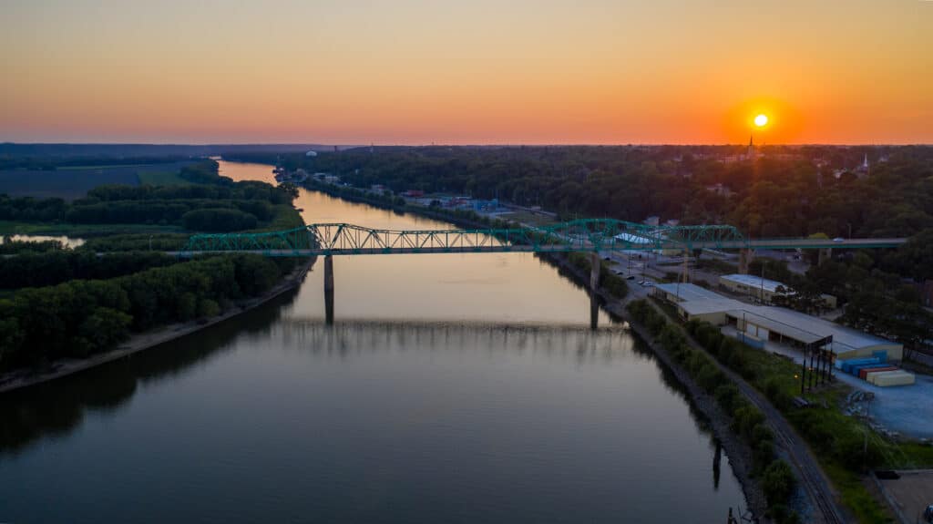 A serene river at sunset reflects the orange glow of the sky. A green truss bridge stretches across the water, with lush greenery lining the banks. The horizon features a silhouetted town skyline against the setting sun.