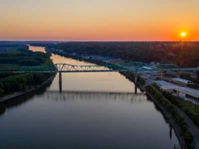 A serene river at sunset reflects the orange glow of the sky. A green truss bridge stretches across the water, with lush greenery lining the banks. The horizon features a silhouetted town skyline against the setting sun.