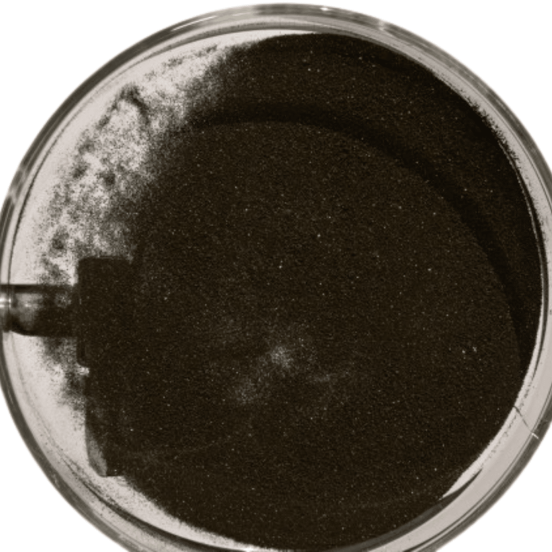 A round, transparent container filled with dark, fine sand sitting against a plain background. The sand appears smooth and evenly distributed, with some slight texture variations visible on the surface.