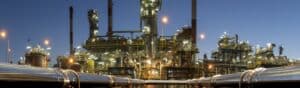 Panoramic view of an industrial oil refinery at twilight. The complex structure is illuminated with bright lights, highlighting various machinery and towering chimneys. In the foreground, metallic pipelines extend towards the refinery.