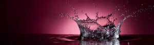 Water splash captured mid-air against a gradient pink background. Droplets and ripples are visible, creating a dynamic, artistic effect.