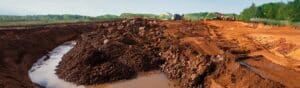 A construction site with large excavation machinery working on a muddy landscape with piles of dirt and a partially dug trench. In the background, green trees and a clear sky are visible.