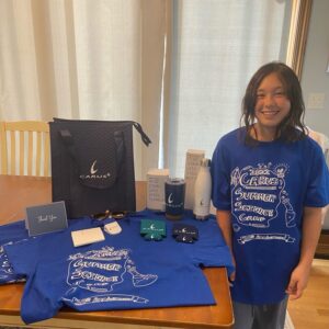 A smiling girl stands next to a table displaying various items, including blue shirts with "Carus Summer Science Camp" design, a stainless steel bottle, a notebook, a bag, and other branded merchandise.