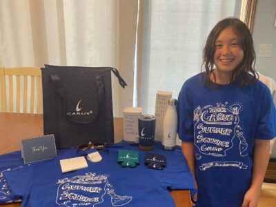 A smiling girl stands next to a table displaying various items, including blue shirts with "Carus Summer Science Camp" design, a stainless steel bottle, a notebook, a bag, and other branded merchandise.