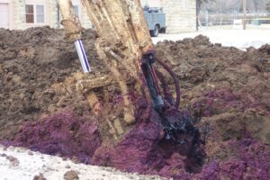 A construction excavator is digging into the ground, revealing vibrant purple soil mixed with dark black material. The background shows a blurred building and a parked truck.