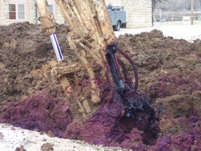 A construction excavator is digging into the ground, revealing vibrant purple soil mixed with dark black material. The background shows a blurred building and a parked truck.