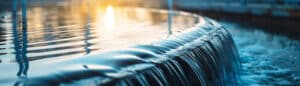 Close-up of water cascading over the edge of a fountain, catching the warm glow of sunlight. The focus is on the smooth flow of water and the shimmering reflections, creating a serene and calming scene.