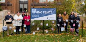 A group of nine people stand in front of a "The Giving Fence" banner with attached bags containing various items. They are outdoors, on a lawn, with a brick building and autumn foliage in the background.