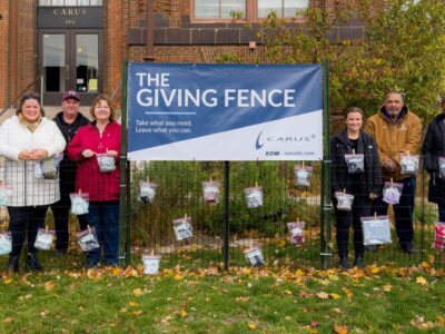 A group of nine people stand in front of a "The Giving Fence" banner with attached bags containing various items. They are outdoors, on a lawn, with a brick building and autumn foliage in the background.