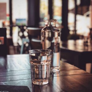 A clear glass of water and a glass bottle set on a wooden table in a dimly lit cafe. The background shows blurred chairs and large windows letting in natural light, creating a cozy atmosphere.