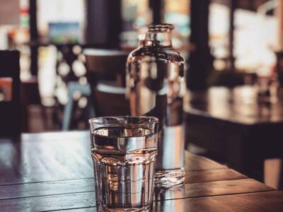 A clear glass of water and a glass bottle set on a wooden table in a dimly lit cafe. The background shows blurred chairs and large windows letting in natural light, creating a cozy atmosphere.